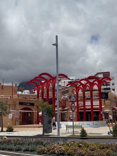 The image shows a street scene in Adeje, Spain, with a large, red, sculptural art installation as the dominant element. The sculpture consists of multiple curved, arching red structures that repeat vertically, creating a striking visual pattern. In the background, there are buildings with beige brick facades and shops, one of which appears to be a bank with a "CaixaBank" sign. Other signs visible include "ITALIAN RESTAURANT," "Mica," and "Perfum," indicating a commercial area.
There are a few people visible in the scene. One person is walking on the sidewalk, and another is standing near a green kiosk or public utility box. A child is also present near a bench. The weather appears overcast, with a cloudy sky. A speed limit sign indicating "30" and a roundabout sign are also present, suggesting a public road. The overall impression is of a moderately busy, urban environment with an artistic flair.