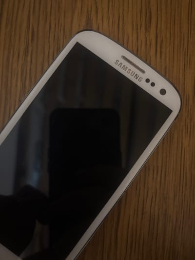 A white Samsung smartphone is placed on a wooden surface. The phone is angled with the top right corner closer to the viewer. The screen is black, indicating it is either off or displaying a dark image. The Samsung logo is visible in silver lettering above the screen, along with the earpiece and two circular buttons. The wooden surface has a prominent grain pattern, oriented vertically. The scene is dimly lit, with a reflection visible on the phone's screen. The location of Радваніцкі сельскі Савет, Belarus, is not discernible from the image.