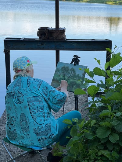 A person is sitting on a chair outdoors, facing away from the camera and towards a body of water. They are painting a landscape on an easel. The person is wearing a light blue shirt with a black tribal pattern of faces and abstract shapes, and turquoise pants. They also have on a light blue and floral patterned cap. The painting on the easel appears to be a forest scene with a tree in the foreground. In the foreground of the image, there is a large, rusty metal object with a screw mechanism, possibly part of an old machine or clamp. To the right of the person, lush green leaves and branches of a tree overhang the scene. The background is a wide expanse of water, reflecting the light of what appears to be late afternoon or early evening. The overall mood is serene and focused, suggesting a peaceful moment of artistic creation in nature. The setting appears to be a rural area near a lake or river, consistent with the provided location context of Пекалінскі сельскі Савет, Belarus. There is no visible text in the image.