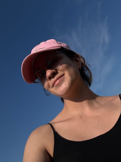 An upward-angle, medium shot captures a young woman wearing a pink baseball cap and round-rimmed sunglasses, looking down at the camera with a slight smile. She is wearing a black tank top. Her hair is brown and appears windswept. The background is a clear blue sky with a few wispy clouds. The image does not provide any visual information to suggest a location in the city of Докша, Russia.