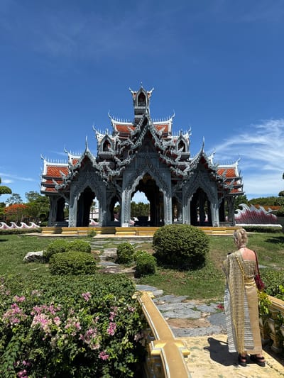 A woman in traditional Thai attire stands next to a highly ornate pavilion. The pavilion features multiple layers of intricate grey architectural details and red tiled roofs under a clear blue sky with some wispy clouds. The surrounding environment consists of meticulously landscaped greenery, including shaped shrubs and flowering plants. The setting suggests a cultural or historical site within the Bang Pu Subdistrict Municipality, Thailand.