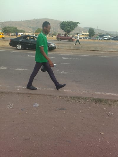 The image shows a man in a green shirt and dark pants walking on the edge of a paved road. He is walking towards the right of the image. The man is holding a black cell phone in his right hand. There is a black car to his left and a red car in the background. There are trees in the background and a hill in the distance. There is a white line painted on the edge of the paved road. There is dirt on the side of the road. There is trash on the ground in the dirt.  The image was taken near coordinates 9.024, 7.573, which is in the city of Maraba, Nigeria.