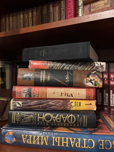 The image displays a static arrangement of books within an indoor environment, specifically identified as Velikiye Luki, Russia. In the foreground, seven books are stacked horizontally, spines facing the viewer. The topmost book has a black hardcover with "Л. ТЕРНИК" (L. TERNIK) visible. Below it is an orange-red book with patterned artwork. The third book is brown, titled "СОБРАНИЕ СОЧИНЕНИЙ ТОМ 1" (COLLECTED WORKS VOLUME 1), featuring an illustration of a quill pen and two figures embracing. The fourth book is red, labeled "МАЙН РИД" (MAYNE REID) with "Всадник без головы" (The Headless Horseman) also visible. The fifth book, with a multi-toned spine, shows "С. ЛЕМ" (S. LEM). The sixth book is black with prominent gold lettering spelling "ФАРАОН" (PHARAOH) and Egyptian-style symbols. The bottom book in the stack is blue, titled "ЗАГАДКИ СТРАН МИРА" (MYSTERIES OF THE COUNTRIES OF THE WORLD) and includes a globe illustration.
In the background, a wooden bookshelf is visible, housing numerous other books arranged vertically. Titles discernible on the upper shelf include "Лондон" (London), "М. Карамзин" (M. Karamzin), "Трагедии" (Tragedies), and "А. Пушкин" (A. Pushkin). To the right, additional books with red and white spines are vertically oriented, some bearing the word "НОВАЯ" (NEW). No people are present, and no active interactions are depicted. The scene is illuminated by ambient light.