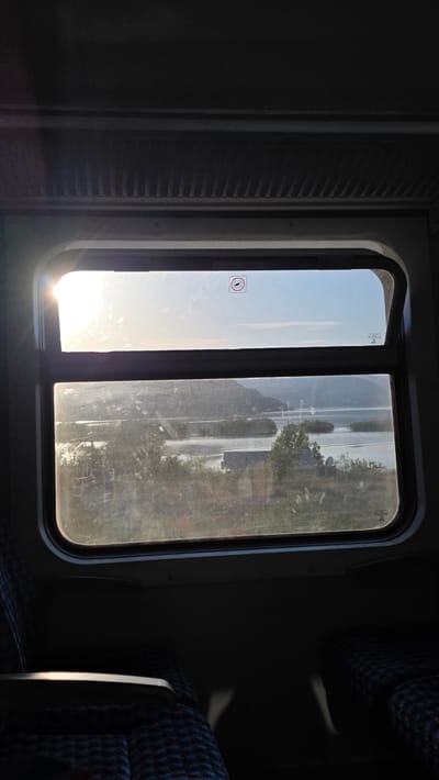 The image presents the interior of a train carriage, focusing on a rectangular window divided into two vertical panes. Strong, low-angled sunlight streams directly into the carriage through the left pane, creating a bright glare and lens flare, suggesting either early morning or late afternoon.
The interior is comparatively dim, with the light from the window casting parts of the carriage into shadow. On the far left, a dark, ribbed panel, possibly a closed window blind or part of the wall, is visible. To the right, a section of a train seat with blue patterned fabric and a silver armrest is seen.
The view outside the window is partially obscured by the intense sunlight, reflections, and visible dirt or scratches on the glass, especially on the right pane. Through the bright haze, a clear sky transitions into a luminous horizon. The distant landscape appears blurred, showing outlines of trees and what might be a road or a body of water, indicating movement through a natural or semi-rural environment.
A small, circular red and white sticker with a crossed-out symbol, likely indicating "do not lean out" or "do not open," is affixed to the left window pane. Faint, translucent symbols or logos, possibly manufacturer marks, are also visible near the top of both panes. The scene evokes a sense of travel through a sunny landscape, potentially in Vranjina, Montenegro.
