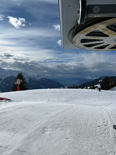 The image depicts a snowy mountain landscape at a ski resort in Villard-sur-Doron, France, viewed from beneath a ski lift mechanism. The upper right portion of the frame shows a large, weathered metal pulley wheel with a patterned surface and a smaller, brighter metal wheel, part of a ski lift's grey housing and cables. The midground and foreground are dominated by a wide, well-groomed ski slope covered in fresh snow, exhibiting distinct parallel corduroy patterns from recent grooming. To the left, a multi-paneled wooden directional signpost, displaying various colored route markers, is mounted on an orange pole beside a tall evergreen tree. To the right, a lone individual, dressed in a white jacket and dark pants, stands on skis, holding ski poles, facing away from the viewer and looking towards the distant terrain. The background features a vast expanse of snow-covered mountains and valleys, with scattered evergreen trees, all under a partly cloudy sky that transitions from bright blue patches to heavier grey cloud formations.