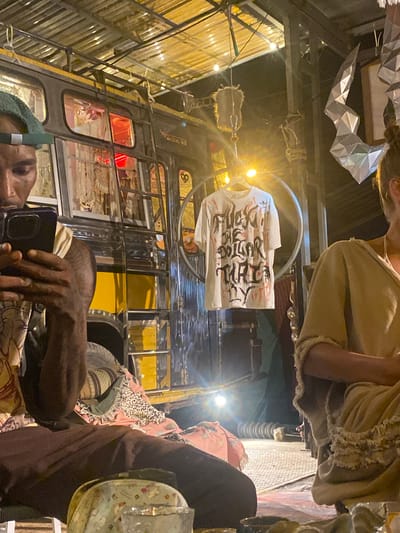 The image, taken in Moo 4, Thailand, shows an unusual composition with the camera tilted upwards. A group of people are visible, with some individuals appearing closer to the lens than others. A person with a green head covering is looking at a phone. A t-shirt with prominent, stylized text hangs in the foreground, suspended from a circular support above a vehicle. The vehicle itself appears to be a converted bus, with yellow and black coloring, and modifications for what looks to be a unique dining or gathering space. The background suggests an outdoor or semi-outdoor venue, with corrugated metal roofing and decorative lighting. The overall ambiance hints at a festive or creative environment.