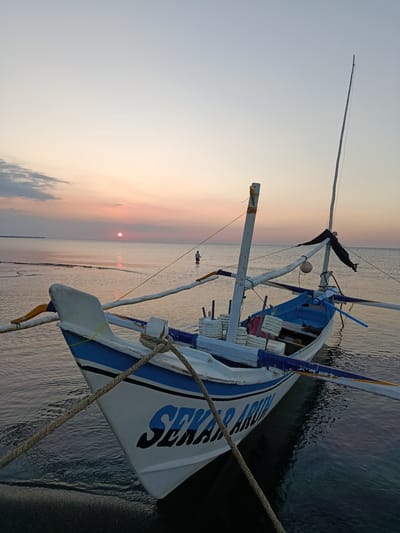 This is a low-angle, full shot photograph of a white and blue fishing boat moored in shallow water at sunset in Jangkar, Indonesia. The boat is tied to the shore with a thick rope and is angled towards the left of the frame. Its name, "SEKAR ARUM," is visible in blue lettering on its bow. The boat has a single mast with rigging and a black flag attached. The water is calm and reflects the soft colors of the sky. In the distance, a lone figure stands in the water, and a pier extends into the sea. The sky is a gradient of pale blue, pink, and orange, with a few wispy clouds. The sun is a small, red orb setting on the horizon, casting a warm glow on the scene. The foreground shows a dark, sandy shoreline.