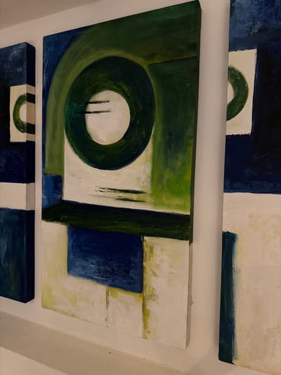 The image shows a close-up, side-angle view of three abstract paintings hanging on a white wall. The paintings are vertical and appear to be part of a series, with a consistent color palette of deep blues, greens, and off-white. The leftmost painting is partially visible, showing a vertical rectangle with a white center and a green crescent shape. The central painting is the most prominent, featuring a large, thick green circle with a white center, bisected by two thin black lines. Below the circle, geometric shapes in blue, green, and white are arranged in layers. The rightmost painting is also partially visible, with a white surface and a vertical dark blue band on the left side. The paintings have a textured, painterly quality. The setting is likely an art gallery or studio, and there are no people or animals present. The overall impression is of modern, minimalist art.