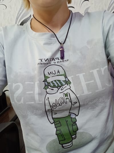 A person is wearing a light blue t-shirt and a purple pendant necklace. The t-shirt has a graphic print of a character wearing a green hoodie and a green and white striped scarf. The character is also wearing glasses and has a peace sign gesture. There is text on the t-shirt, but it is partially obscured and difficult to read. The person's neck and shoulder are visible, and their hair is blonde. The background appears to be a bed with a patterned comforter. The lighting suggests it is daytime. The overall mood is casual and relaxed.
Visible text:
- "DESIGN" (reversed)
- "PRINT" (reversed)
- "KM"
- "HOHO" (partially visible)
- "2" (partially visible)
- "1-" (partially visible)