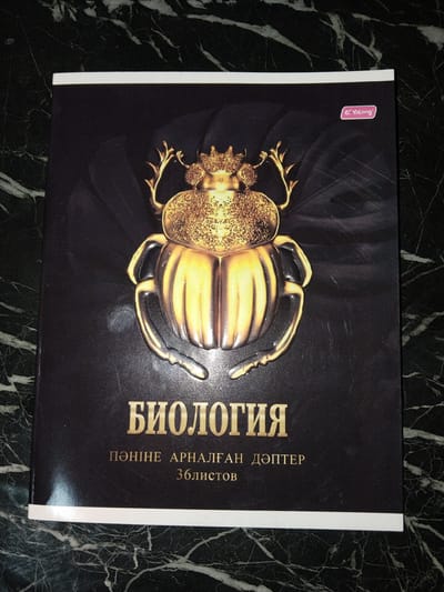 The image shows the cover of a notebook with the title "БИОЛОГИЯ" (BIOLOGY) printed in gold lettering. Below the title, there is text in a smaller font that appears to be in the Kazakh language. The cover features a large, ornate golden scarab beetle design in the center. In the upper right corner, there is a pink oval logo with the word "Yalong" inside. The notebook is placed on a dark green, veined surface that resembles marble. The overall impression is that of a school notebook or textbook cover. There are no people or actions depicted in the image. The location in Ақадыр, Kazakhstan is not discernible from the image itself, as it only shows the notebook cover and the surface it rests on.