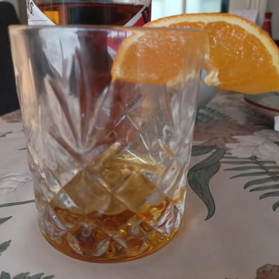 A close-up shot captures a glass of amber liquid, likely whiskey, with an orange slice perched on its rim. The glass has a cut-glass pattern, and the liquid is illuminated by an unseen light source, creating a warm glow. In the background, partially obscured by the glass, is a bottle with a label that reads "B.S." and "TURB." The scene appears to be indoors, set on a table with a floral patterned tablecloth. The overall impression is one of a casual moment, perhaps an evening drink. The lighting suggests it could be anytime, but the ambiance leans towards the evening. No specific weather conditions are discernible. There are no people visible in the image.