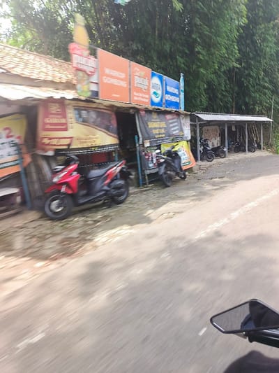 The image shows a street in Sukasari, Indonesia, at coordinates -6.837 latitude and 107.598 longitude. The street is paved with asphalt and lined with a small building on the left side and a dense bamboo forest on the right. The building has several signs, including advertisements for a water brand, "Warung Gembong". The building also appears to be a small shop or store. There are several motorcycles parked in front of the building and under a metal awning. The photo is taken from the perspective of a person riding a motorcycle on the street. The person is looking ahead and the front mirror of their motorcycle is visible in the lower right corner of the image. The motorcycles are parked facing a wall.  The ground is a light gray concrete. The day is bright and the bamboo leaves are green. There are no people visible in the image.