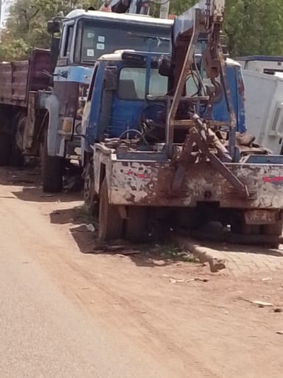 The image shows two large, worn-down trucks parked on a dirt road in Dungu, Ghana. The first truck is a flatbed type, while the second is a blue tow truck. The paint on both vehicles is faded and chipped, with visible signs of rust. The background includes sparse trees and a glimpse of a white van. The ground is dry and dusty. No people are visible in the image. The tow truck's crane mechanism is prominently displayed.