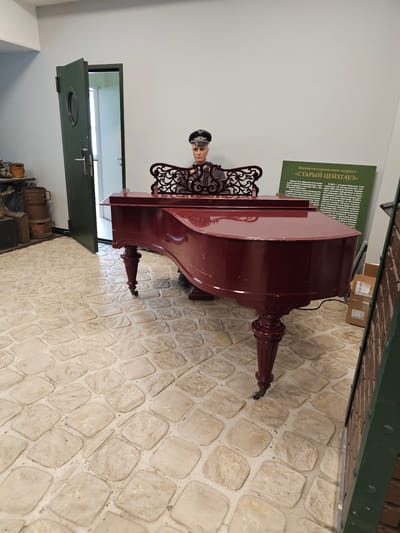 The image captures a scene within a building, potentially a museum or historical site, possibly in Архангельское, Russia. A reddish-brown grand piano dominates the scene, placed on a floor with light-colored stone tiles. Behind the piano is a lifelike mannequin dressed in what appears to be a historical military or official uniform, positioned as if playing the instrument.
A green sign with Russian text is mounted on the wall near the piano, likely offering information or context about the display. To the left, a partially open dark green door leads to another room with a glimpse of a white interior and window. Below the doorway, a collection of vintage suitcases and trunks are arranged.
The room's decor and the presence of the mannequin and informational sign suggest a staged exhibit or historical recreation.