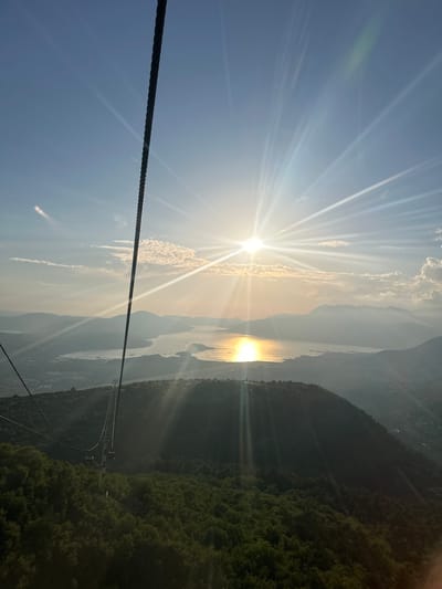 The image shows a view from a cable car ascending Mount Vrmac overlooking the Bay of Kotor near Mirac, Montenegro. The sun is shining brightly, creating lens flares and a reflective path on the water. The cable car's thick rope is visible in the foreground, with a cable car carriage descending on a separate line. Lush green trees cover the steep slopes of Mount Vrmac, with a small town visible in the distance along the coast. The bay is surrounded by hazy mountains, suggesting a scenic and elevated perspective.