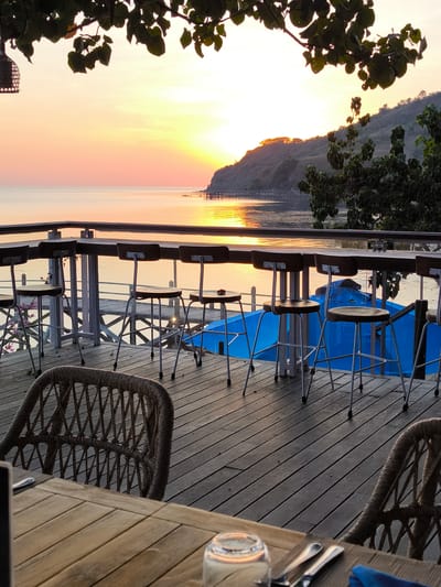 A view of a sunset over the ocean from a deck with chairs and tables. The deck is made of wooden planks and overlooks the water. There are several high chairs with brown seats and metal legs lined up against a wooden bar. In the foreground, a woven wicker chair and a wooden table with place settings are visible. The ocean reflects the orange and yellow hues of the sunset. In the distance, a hilly coastline is visible, dotted with trees. The sky is a gradient of orange, pink, and yellow, with a few clouds. The scene is set at a restaurant or bar in Labuan Bajo, Indonesia, likely on the waterfront.