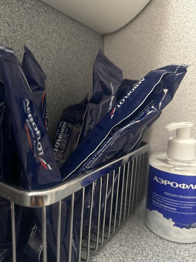 The image showcases several Aeroflot-branded toothbrush and toothpaste packets arranged in a chrome wire rack. A bottle of Aeroflot lotion is also visible in the top right. The packets are dark blue with white lettering. The lotion bottle is white with blue text, stating that it is a moisturizing lotion with regenerating properties. The packets contain a toothbrush and toothpaste. The composition details the toothpaste's ingredients and manufacturing.