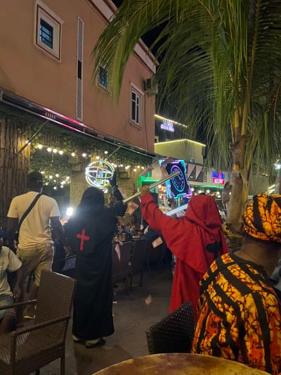 The image depicts an outdoor nighttime scene at what appears to be a resort or hotel in Oba, Nigeria. In the foreground, two individuals are the primary focus. One person is wearing a black hooded robe with a red cross emblem on the back and is holding up a metallic pole. To their right, another person is wearing a red hooded robe and also appears to be holding a metallic pole, possibly in interaction with the first person. Further to the right, another individual is visible from the shoulders up, wearing a vibrant orange and black patterned headwrap and garment.

The background reveals a building with illuminated signage, including "LUFTGA HOTEL & SUITES" and another sign that reads "RESORT." There are tables and chairs scattered throughout the area, suggesting an outdoor seating or dining space. Palm trees with lush green fronds are visible, and decorative lights are strung along an overhead structure, illuminating the scene. The ground appears wet, indicating recent rain or a damp surface. The overall atmosphere suggests a gathering or event taking place at this location in Oba, Nigeria.