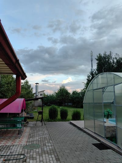 The image depicts a backyard scene at dusk in the Фаніпальскі сельскі Савет, Belarus. In the foreground, a paved walkway made of grey and pink interlocking bricks leads towards a large, transparent greenhouse on the right. Inside the greenhouse, a white vase with a green artificial plant is visible next to a body of water. To the left of the walkway, a covered seating area with green benches and a table is visible under a red-tiled roof and pink canopy. A metal barbecue grill stands next to the seating area. In the background, a two-story house with a grey roof and windows is partially visible behind a metal chimney. Dense green trees surround the property. A tall cell tower with antennae is visible in the distance, silhouetted against a cloudy sky with hints of pink and orange from the setting sun. The overall environment suggests a rural or suburban residential setting. There are no people or animals visible in the image.