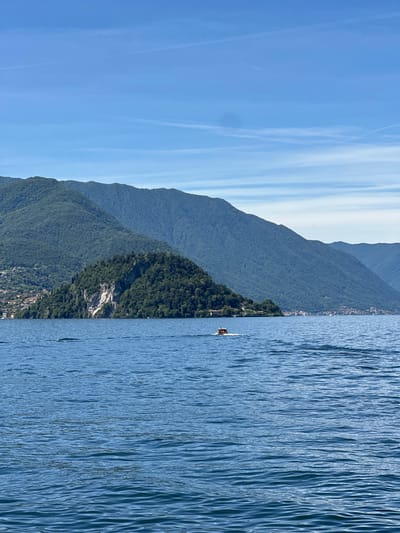 A wide-angle, eye-level shot captures a serene lake scene with a vibrant blue sky. In the foreground, the calm, rippling surface of the lake dominates the lower half of the frame. A small, red and white boat is visible in the middle ground, moving from left to right across the water, leaving a white wake.
The background features a lush, green mountainside that rises steeply from the lake's edge. The slopes are densely covered with trees, with hints of scattered buildings and homes visible along the shoreline on the left. The mountains extend into the distance, with layers of progressively darker blue peaks creating a sense of depth. The sky is a clear, bright blue with a few wispy, white clouds streaking across it, likely contrails from aircraft. The overall atmosphere is peaceful and picturesque, evoking a sense of summer tranquility.