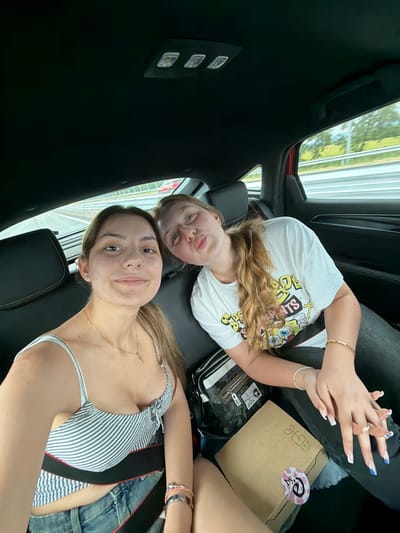 Two young women are inside a vehicle, possibly a car. The first woman, closest to the camera, is wearing a striped top and jeans with a visible seatbelt. She has light brown hair and a necklace. The second woman, behind her, is wearing a white t-shirt with a graphic design. She has blonde hair. Their hands are clasped together. Several items are on the seat, including a black and white patterned bag and a brown box with a sticker. The background shows a blurred view of trees and structures outside the car window. There is no indication that the image was taken in Chokoba, Bulgaria.