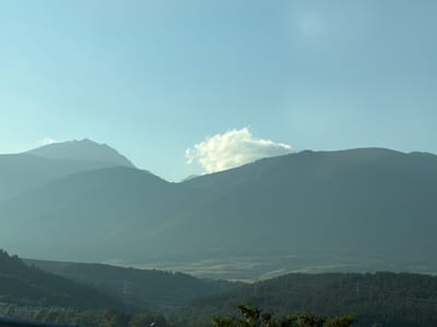 The image depicts a panoramic view of mountains and a valley, likely taken from an elevated position. The sky is a clear, light blue with a few wispy white clouds, one particularly prominent cloud cluster situated above the central mountain range. The mountains themselves are layered, with the closest ones appearing a deep, muted green, transitioning to lighter, hazy blues and grays in the distance, suggesting depth and atmospheric perspective. The foreground shows a wooded slope with a hint of a road or barrier at the very bottom. There are no people or animals visible. The overall impression is a natural landscape, possibly captured at dawn or dusk due to the soft lighting and muted colors. This scene could be a representation of the landscape surrounding the city of Djerman, Bulgaria, known for its mountainous terrain.