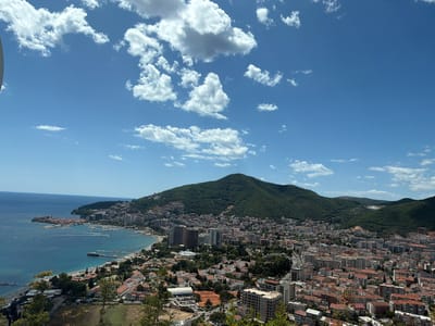 This is a high-angle, wide shot of the coastal city of Budva, Montenegro, with the island of Košljun visible in the distance on the left. The sky is a vibrant blue with scattered white cumulus clouds. The sea is a deep blue, meeting a rocky coastline dotted with buildings. The city itself is a dense collection of residential and commercial buildings with red-tiled roofs, nestled against a lush green mountainous backdrop. A prominent green hill with a tower on its summit dominates the central and right portions of the image. The foreground consists of trees and foliage, suggesting the viewpoint is from a higher elevation overlooking the city. A beach area with umbrellas and a pier is visible along the coastline, and several boats are anchored in the bay. There are no people or animals clearly discernible in the image. The overall scene depicts a sunny day in a Mediterranean coastal city.