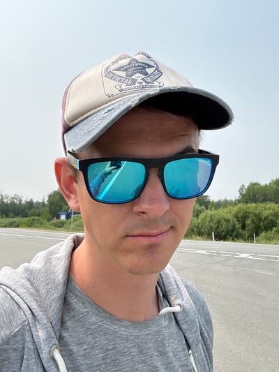 A man wearing sunglasses and a baseball cap is taking a selfie outdoors. He is wearing a grey hooded jacket over a grey t-shirt. The background shows a road and trees, suggesting a rural or suburban setting. There is a hint of a building in the distance. The image does not contain enough information to confirm the location as Kargat, Russia.