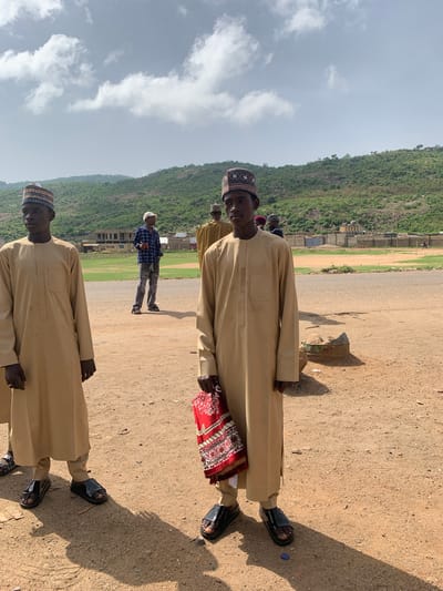 The image depicts several men in a public space in Naraguta Hausawa, Nigeria. The men are wearing traditional garments, including caps and long robes. One man holds a red prayer mat. They stand on a dirt ground. A building under construction is visible in the background. A green, hilly landscape is also present. The sky is partly cloudy.