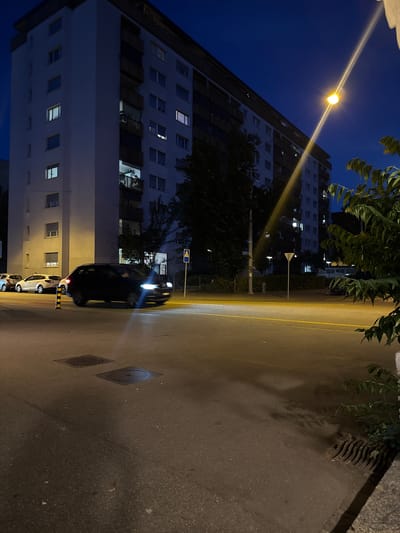 The image depicts a nighttime street scene in Basel, Switzerland. A tall residential building with numerous windows, some illuminated, dominates the left side. A street light is visible, casting a bright beam onto the building and the street. A black car with its headlights on is driving on the right side of the street, which has a yellow center line. Several white parked cars are visible in the background. Trees and other foliage are present along the street, obscuring some of the building. A pedestrian crossing sign is visible. Two square metal road covers are on the street. The sky is a dark blue.