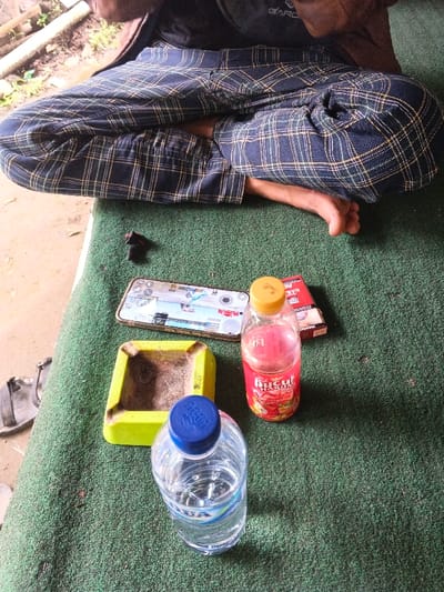 A person sits cross-legged on a green surface. A smartphone displays a game, positioned next to a yellow ashtray, a red juice bottle, and a water bottle. A pack of cigarettes and a dark object sit nearby. The person wears plaid pants. One foot is visible.