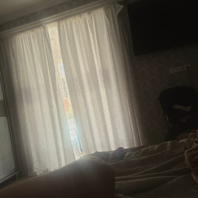 An indoor scene captures a view of a bedroom with soft, diffused natural light coming through sheer white curtains. The curtains are patterned with a delicate, swirling motif and are drawn to reveal a glimpse of the outside, which includes a wrought-iron window grate and possibly some colorful objects beyond. To the right of the curtains, a dark, large flat-screen television is mounted on a wall. Below the television, a wall features patterned wallpaper and a white electrical outlet with two sockets. A dark, bulky object, possibly a chair with clothing draped over it, is visible on the right side of the frame. In the foreground, a person's legs and feet are visible, lying on a rumpled, beige blanket or duvet. The overall impression is of a cozy, lived-in bedroom, likely in a residential setting within Zheleznovodsk, Russia, suggested by the provided context.