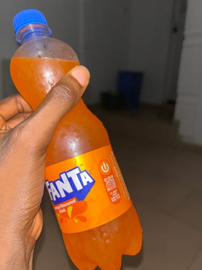 The scene displays a dark-skinned hand holding a clear plastic bottle of Fanta orange-flavored drink. The bottle, covered in condensation, features a blue screw cap and an orange label with the "FANTA" logo in blue and white, "orange flavoured drink" text, a QR code, and the slogan "SIP, SHAKE & SCAN TO UNLOCK THE FUN." The environment is an indoor location in Egbe, Nigeria, characterized by low ambient lighting. The background is dark and out of focus, showing a blurred floor and a distant, dark blue object, consistent with a container.