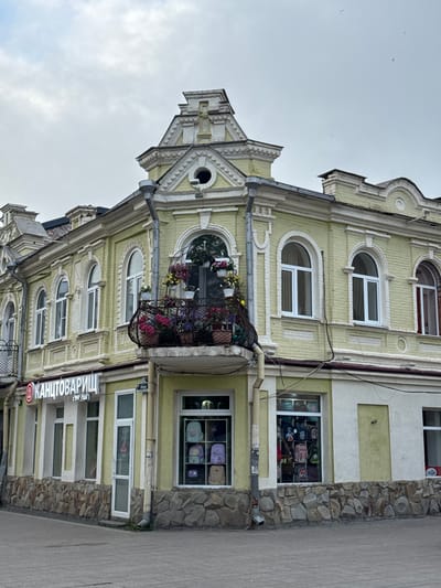 The image depicts a multi-story building in Akhieli, Georgia. The building features an ornate facade with arched windows, a small balcony adorned with flowers, and a decorative gable. The exterior is painted in light yellow and white. A sign in Cyrillic script, “Канцтоварищ (since 1997 years),” indicates a stationery store at the ground level. The windows on the ground floor display merchandise, including backpacks. The building is situated alongside a paved street. The sky is overcast.