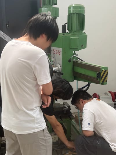 The image depicts three young men, likely students, gathered around a green milling machine in what appears to be a workshop or laboratory setting. One man, wearing a white t-shirt and grey pants, stands to the left, his back mostly to the camera, looking down at the machine. Another man, also in a white t-shirt and dark pants, is crouched down to the right, actively working on the machine. He is wearing glasses. A third man, seen from the side and slightly from behind, is wearing a black t-shirt with white sleeves and is also engaged with the machine. The milling machine is a prominent feature, colored bright green with various controls and a digital display showing "23032". The environment suggests an industrial or educational space with white walls and possibly other equipment visible in the background. The men are focused on their tasks, indicating a learning or practical application session. The specific location in Shuangfu, China, cannot be determined from the visual information presented in the image.