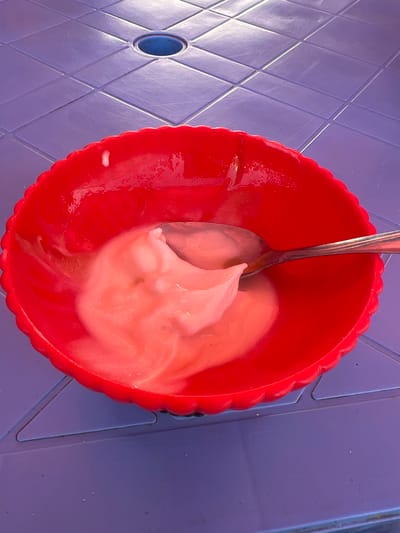 A red, scalloped bowl filled with a pink, creamy substance, possibly yogurt, rests on a blue plastic table. A silver spoon is partially submerged in the substance. The table has a grid pattern and a circular hole in the center. The scene is outdoors, with sunlight illuminating the food and table. The image was taken in the city of Balkh, Tajikistan.