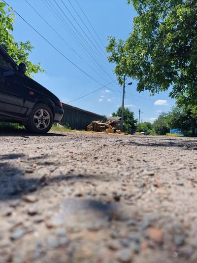 A low-angle, eye-level shot captures a rural road in Bershad, Ukraine, under a clear blue sky dotted with fluffy white clouds. The foreground is dominated by the rough texture of the unpaved road, with scattered pebbles and dirt. To the left, the dark body of a car is partially visible, its wheel and side in sharp focus. Behind the car, a long, weathered green fence runs horizontally across the frame. A pile of cut logs rests against the fence, suggesting recent activity.
Further down the road, a wooden utility pole with wires stretches into the distance, accompanied by a street lamp. Trees with lush green foliage line the sides of the road, casting dappled shadows. In the background, other buildings and more trees can be seen, with a hint of yellow wildflowers blooming near the road. The overall impression is one of a quiet, sun-drenched day in a rural Ukrainian setting.