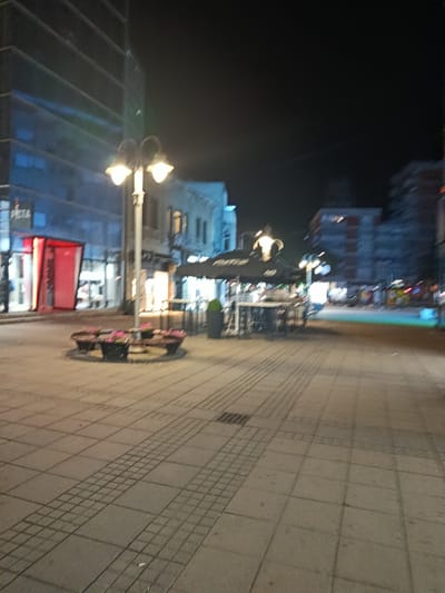 The image depicts a street scene at night in the city of Niš, Serbia. In the foreground, a paved pedestrian walkway with a patterned tile design stretches into the distance. A circular planter with pink flowers sits to the left of the walkway. A street lamp with two lights stands near the planter. Beyond the walkway, a row of buildings lines the street. To the left, a modern building with horizontal windows is illuminated. Further down the street, a building with awnings and outdoor seating suggests a cafe or restaurant. In the background, other buildings of varying architectural styles are visible, some with illuminated windows. The overall atmosphere is dimly lit, with the glow of streetlights and building interiors creating a nighttime ambiance.