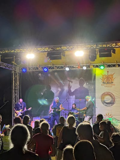 This image depicts a lively outdoor concert scene at night, likely during a summer festival. A band is performing on a brightly lit stage, with multiple spotlights and stage lights illuminating the performers and the backdrop. The band members, dressed casually, are playing guitars and appear engaged in their music. In the foreground, a crowd of people is visible, their backs to the camera, watching the performance. The audience members are silhouetted against the stage lights, some with their heads bowed, indicating focused attention. The overall atmosphere appears energetic and celebratory.
A large banner serves as the stage backdrop, featuring graphics and text in both English and Cyrillic. The English text clearly reads "JULY MORNING TUTRAKAN," suggesting the name and location of the event. The Cyrillic text, which includes "ОГНЕНИЯТ ДУНАВ" (Ogneniyat Dunav, meaning "The Fiery Danube") and "ТУТРАКАН" (Tutrakan), further confirms the location and possibly the theme of the festival. The presence of a stylized sun graphic with a guitar suggests a rock or folk music genre. The time of day is clearly nighttime, indicated by the dark sky and the prominent stage lighting. The weather appears to be clear, with no visible rain or adverse conditions. The scene conveys a sense of community and shared enjoyment of live music.