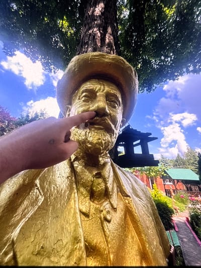 A low-angle, medium shot shows a golden statue of a man wearing a hat and tie. The statue's nose is being poked by a person's finger, which has a small heart tattoo on the back of the hand. The statue is situated outdoors, with a large tree and lush green foliage in the background. In the mid-ground, a wooden birdhouse is visible, along with a structure with a green roof. The sky above is blue with scattered white clouds. The scene is set in the city of Vityazevo, Russia.