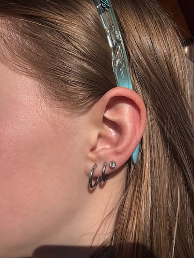 A close-up photograph shows a person's left ear with multiple piercings. The uppermost piercing is a small stud with a clear gemstone. Below it are two silver-colored hoop earrings, creating a stacked effect. The ear is pinkish in color, contrasting with the silver jewelry. The person's light brown hair is visible around the ear. A light blue hair clip with illegible silver characters is partially visible behind the ear. The skin tone appears fair and without blemishes. The background is out of focus, suggesting a shallow depth of field. No elements suggest a location within Gory, Russia.