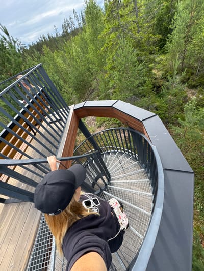 An outdoor, eye-level, high-angle shot shows a woman on a spiral staircase in a forest. The woman is wearing a black baseball cap, a black hooded sweatshirt, and sunglasses perched on her head. She is holding onto the railing of the staircase with her left hand, and her right arm is extended, likely holding a phone or camera. Her hair is blonde and falls over her shoulders. A patterned bag is slung over her shoulder. Behind her, another person is visible on a wooden platform, wearing a camouflage-patterned shirt and holding a piece of paper. The spiral staircase is made of metal with a grated surface. The surrounding environment is a dense forest with numerous green trees and some undergrowth. The sky above is partly cloudy. The image does not provide any specific location details about Peski, Russia, but depicts a natural, wooded setting.