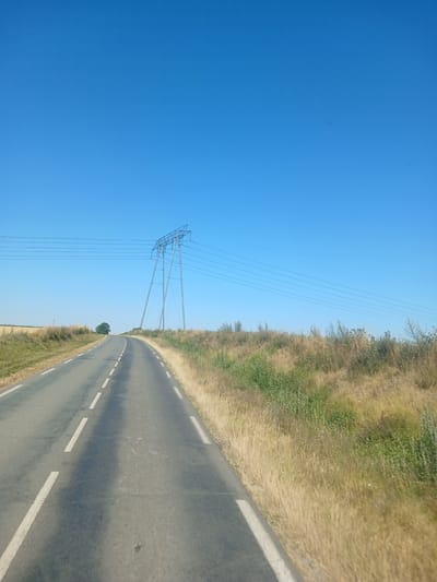 The image captures a rural landscape under a clear blue sky. A paved road with a dashed white line marking the center curves to the left, bordered by dry, golden grass and sparse green vegetation. In the distance, a large, modern electricity pylon with multiple high-voltage lines stands prominently on the right side of the road. A lone tree is visible on the left side of the road, further in the distance, in front of a field of dry crops. The scene appears to be a snapshot from a moving vehicle, suggested by the perspective and the slight motion blur on the road. The overall impression is one of a serene, open countryside in Cléry-sur-Somme, France, characterized by agricultural land and infrastructure.