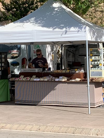 The image shows a market stall with a white tent, set up on a paved area in front of a building. A man wearing sunglasses and a dark t-shirt is standing behind a counter, arranging items. The counter displays various packaged goods, possibly food items, with some wrapped in plastic and others in small boxes. Behind the man, a white van is visible, with its back doors open, revealing storage shelves and some hanging items. To the right of the van, a refrigerated display case contains several white cups with yellow flowers. The market stall has a striped fabric covering the lower portion of the counter. The background includes greenery from trees and a textured wall. The image appears to be taken in Collbató, Spain, as indicated by the context of the provided crops.