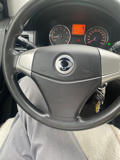 The image shows a close-up view of the interior of a car, specifically the steering wheel and dashboard. The steering wheel has a black leather grip and silver accents. In the center of the steering wheel is a circular emblem with a stylized logo. Below the emblem, "SRS AIRBAG" is visible. To the left of the steering wheel, a turn signal stalk with controls for lights is partially visible. On the dashboard, a digital display shows "OUTSIDE 20" and "40" with some graphics, next to a speedometer that reads up to 220 km/h. To the right of the speedometer is another stalk, likely for windshield wipers, with settings like "MIST," "OFF," "AUTO," and "SLOW." A set of car keys with a fob labeled "CRUISE ON-OFF," "ACCEL ▲," and "DECEL ▼" is attached to the steering column. The lower part of the image shows a person's legs covered in gray pants, and a white fluffy seat cover. The scene is lit by the car's interior lighting, with an orange glow from the dashboard display. No information about the city of Gavrilovka, Russia, can be derived from the image.