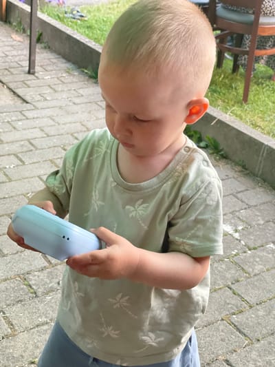 A young boy with blonde hair, wearing a light green t-shirt with a palm tree pattern and blue pants, stands on a paved walkway in Beltershain, Germany. He is holding a light blue object in his hands and looking down at it. The walkway is made of grey interlocking pavers. In the background, there is green grass and a hint of outdoor seating, suggesting a backyard or garden setting. The lighting is bright, indicating it is daytime.
