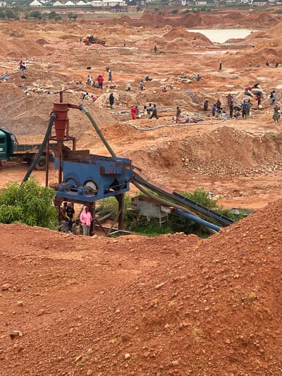 The image depicts an open-air mine, likely a gold mine, in Reifild-Ist, Nigeria. The landscape is characterized by large, excavated pits and mounds of reddish-brown earth. Several individuals, appearing to be workers, are engaged in mining activities throughout the scene. Some are seen carrying loads on their heads or shoulders, while others are gathered around processing equipment.
In the foreground, a blue processing machine is prominent, with a red hopper on top and conveyor belts extending from it. A green truck is parked to the left of the machine. Various hoses and pipes are visible, suggesting the presence of water being used in the mining process. The background shows more distant pits, a body of water, and a settlement with white-roofed buildings. The overall impression is one of active, labor-intensive mining operations in a rural or semi-rural setting within Nigeria.