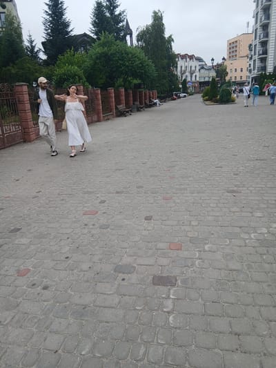 The image shows a street scene in Truskavets, Ukraine. Two people are walking on a cobblestone path. The person in front is a woman wearing a white dress and light pink shoes. The person behind her is a man wearing a white t-shirt, light-colored pants, a dark jacket, a baseball cap, and dark shoes. They appear to be walking away from the camera, towards a row of buildings and lush green trees.

The background includes a multi-story building with balconies and arched windows, as well as other buildings with red-tiled roofs. The sky is overcast, suggesting a cloudy day. The cobblestone pavement is made of grey rectangular stones, with some red and dark grey stones interspersed. A decorative metal fence with intricate scrollwork lines the left side of the path.

There are no visible signs with text. The overall atmosphere appears to be leisurely, possibly a stroll through a town or resort area. The subjects' postures suggest they are engaged in conversation or simply enjoying a walk. The time of day is likely daytime, given the brightness, but the overcast sky prevents strong shadows.