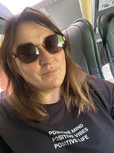 A woman wearing sunglasses and a black t-shirt with white text that reads "POSITIVE MIND POSITIVE VIBES POSITIVE LIFE" is taking a selfie on a bus. She has shoulder-length brown hair. The bus interior is visible, with dark grey seats and a yellow overhead compartment. The location is identified as a bus in Ivanovka, Russia.