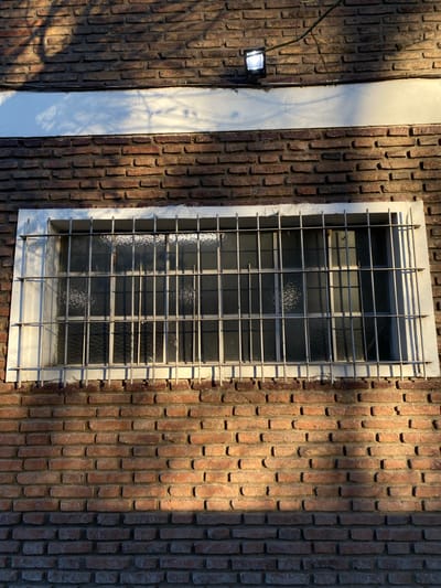 A ground-level, medium shot depicts an exterior brick wall with a barred window. The brick wall consists of red bricks. To the left of the window is a white vertical column, also part of the building's facade. A light fixture is visible on the left side of the column. Shadows from trees and branches are cast across the brick wall and column. The window has a white frame and a metal security grill over the glass. The image was taken in El Talar, Argentina.